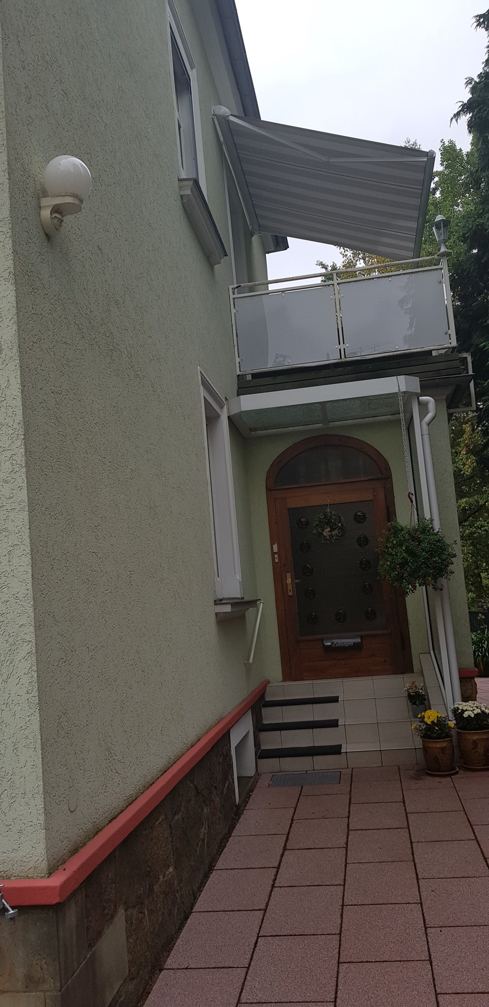 Seiteneingang mit gewölbter Holztür, Stufen und Blumentöpfen, darüber ein Balkon mit Markise