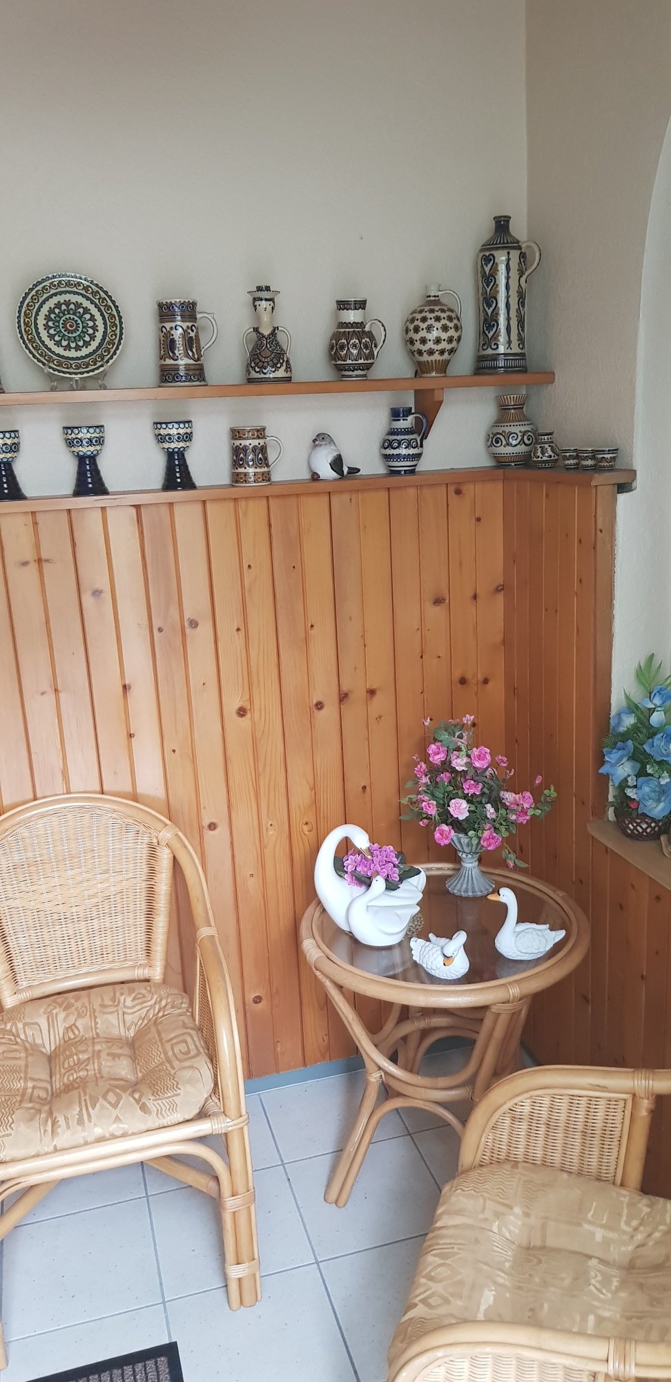 Sitzecke mit Korbsesseln, Glastisch und Holzverkleidung, darüber ein Regal mit Keramikkrügen